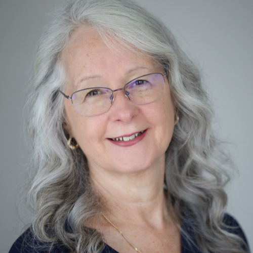 A white woman with grey hair and glasses is wearing a dark shirt and a gold necklace.